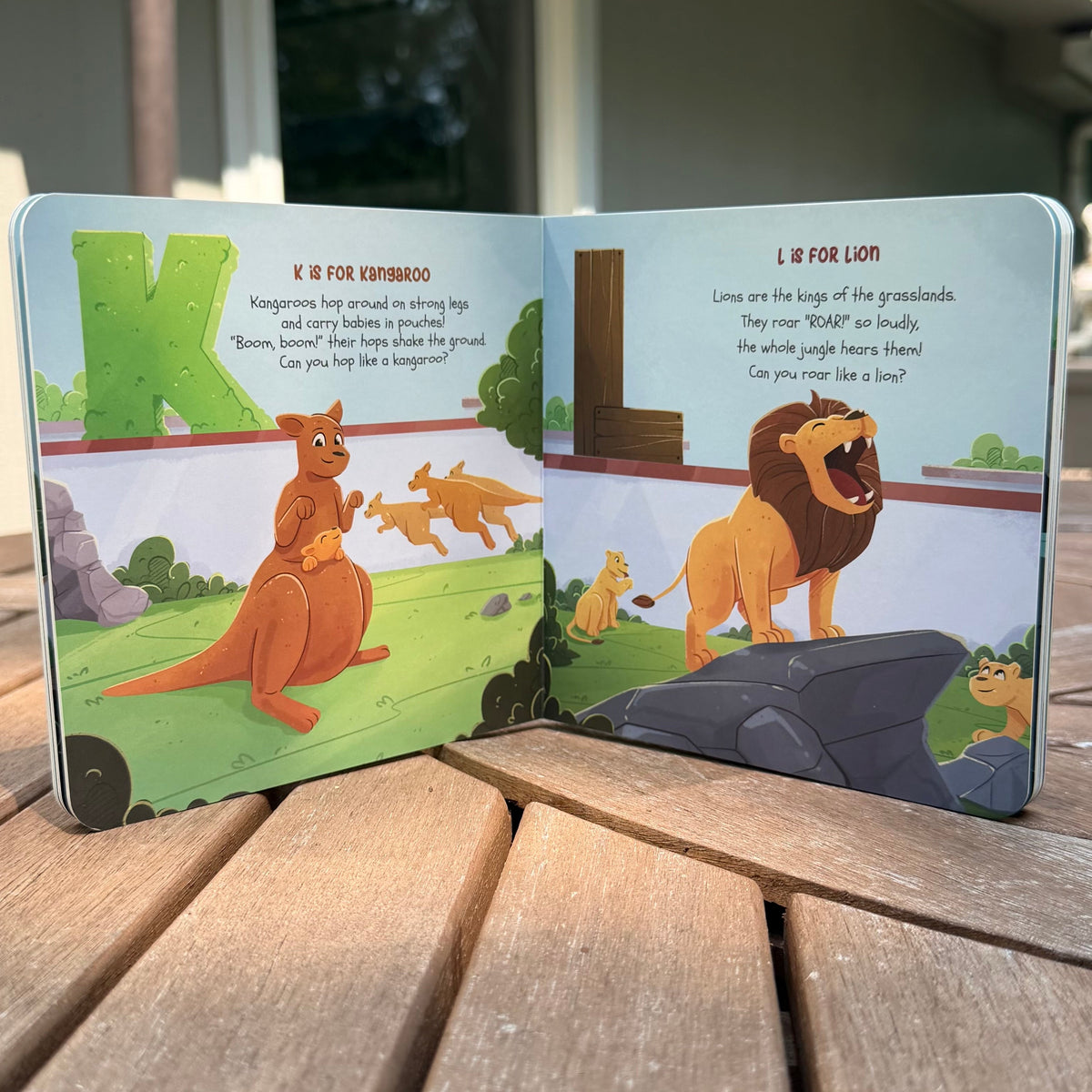 An open children's picture book with colorful animal illustrations and text, photographed standing on a wooden deck outdoors near a house.